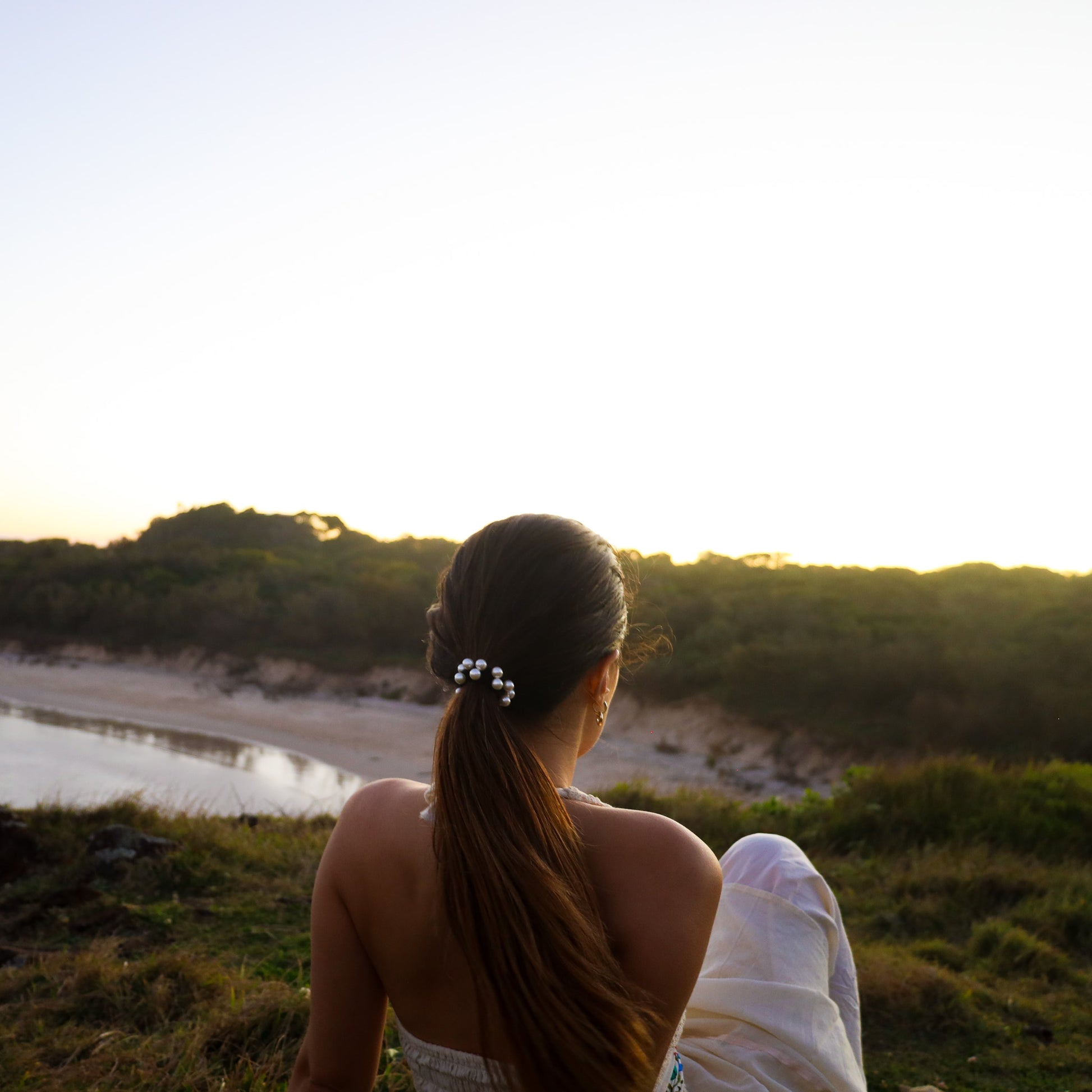 Woman sitting by the ocean at sunset, facing away from the camera.